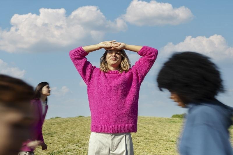 jeune fille au pull rose, debout, portant les deux mains au front et parmi un groupe de jeunes en mouvement