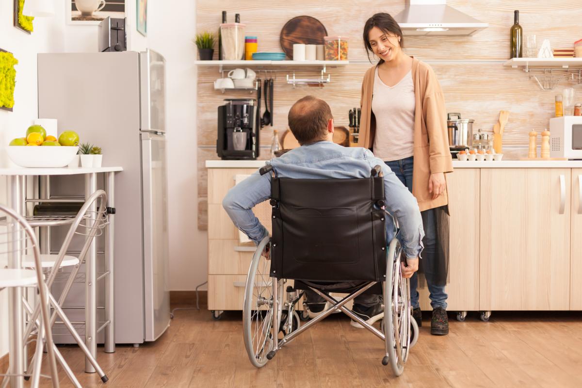De face, femme jeune, souriante qui regarde un homme de dos assis dans un fauteuil roulant. Ils sont dans une cuisine qui paraît adaptée et moderne